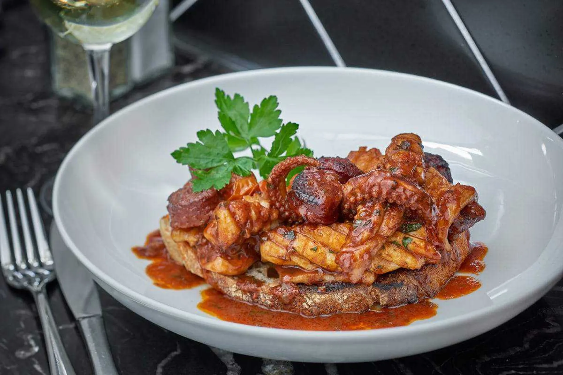 Plated gourmet dish with seared scallops on red sauce, garnished with fresh parsley, served with white wine on dark table setting.