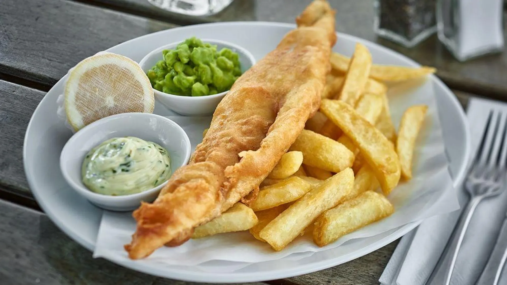 Classic fish and chips meal served on white plate with tartare sauce, mushy peas, and lemon wedge, accompanied by cutlery on wooden table