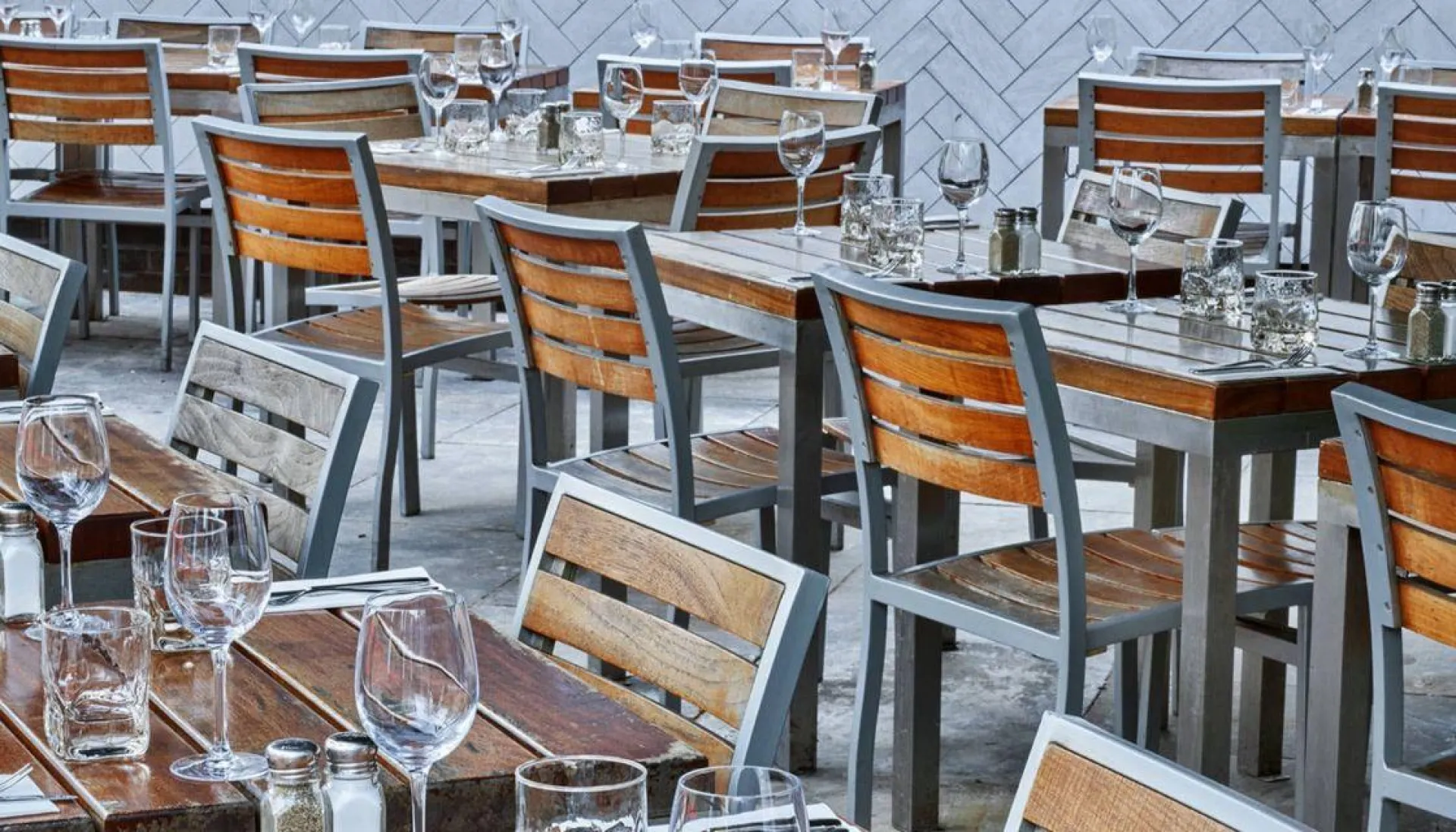 Outdoor dining area with wooden chairs featuring metal frames arranged around tables set with glasses and place settings on a patterned floor.