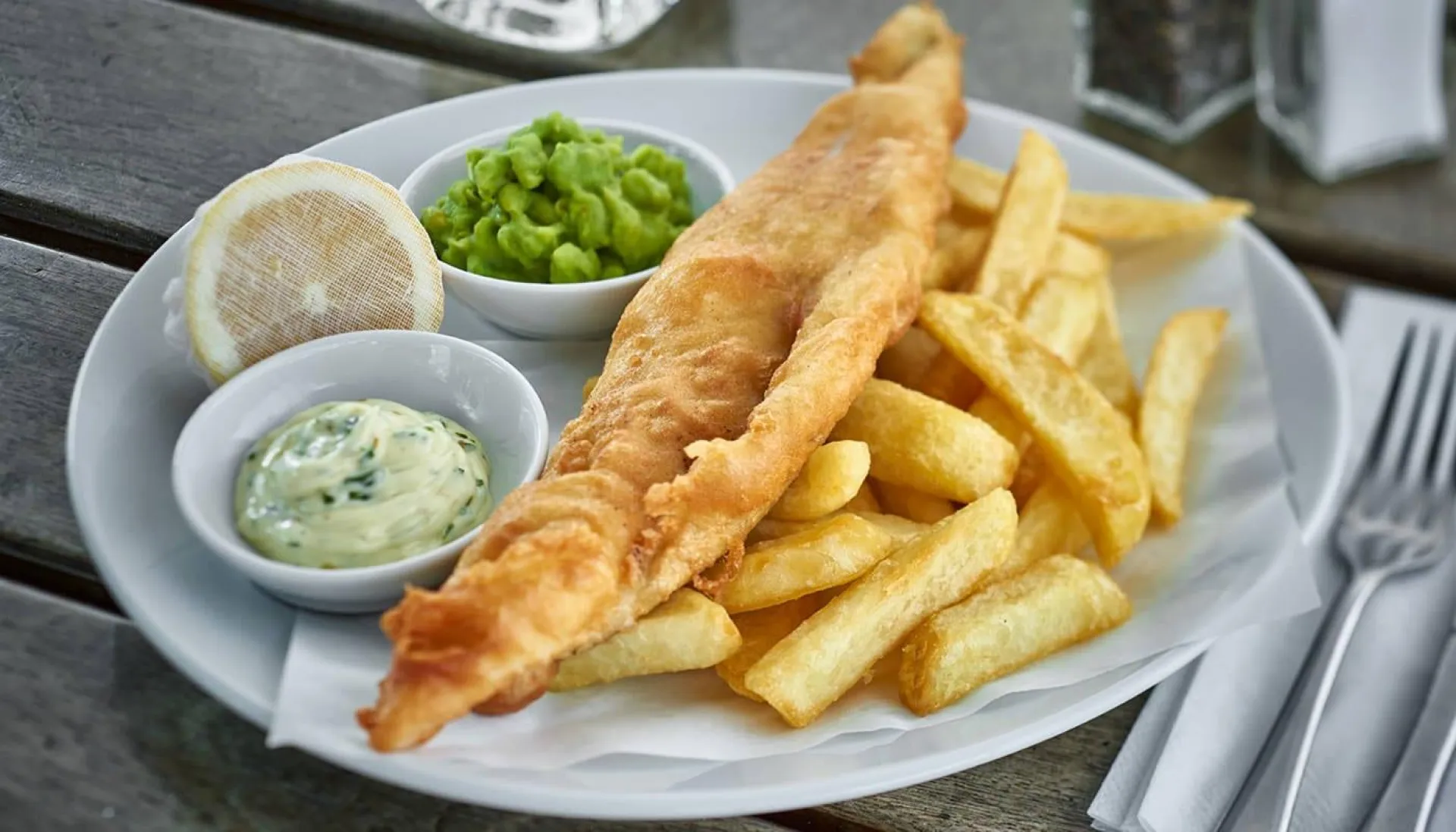 Fish and chips meal served on a white plate with tartare sauce, mushy peas, and lemon wedge, accompanied by cutlery on a wooden table