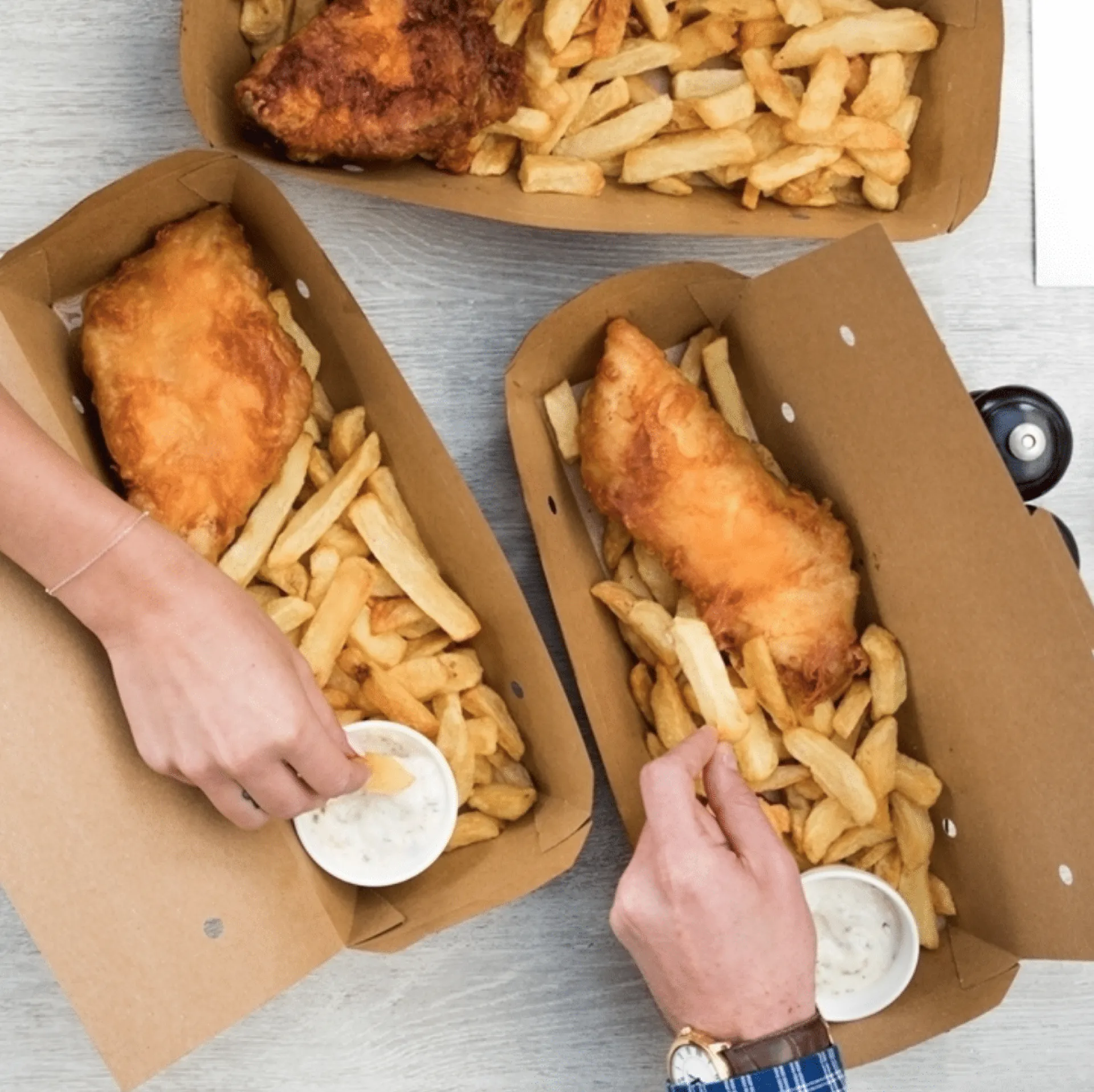 Three cardboard takeaway boxes containing battered fish and chips with white sauce cups, served on a wooden surface with hands reaching for the food.