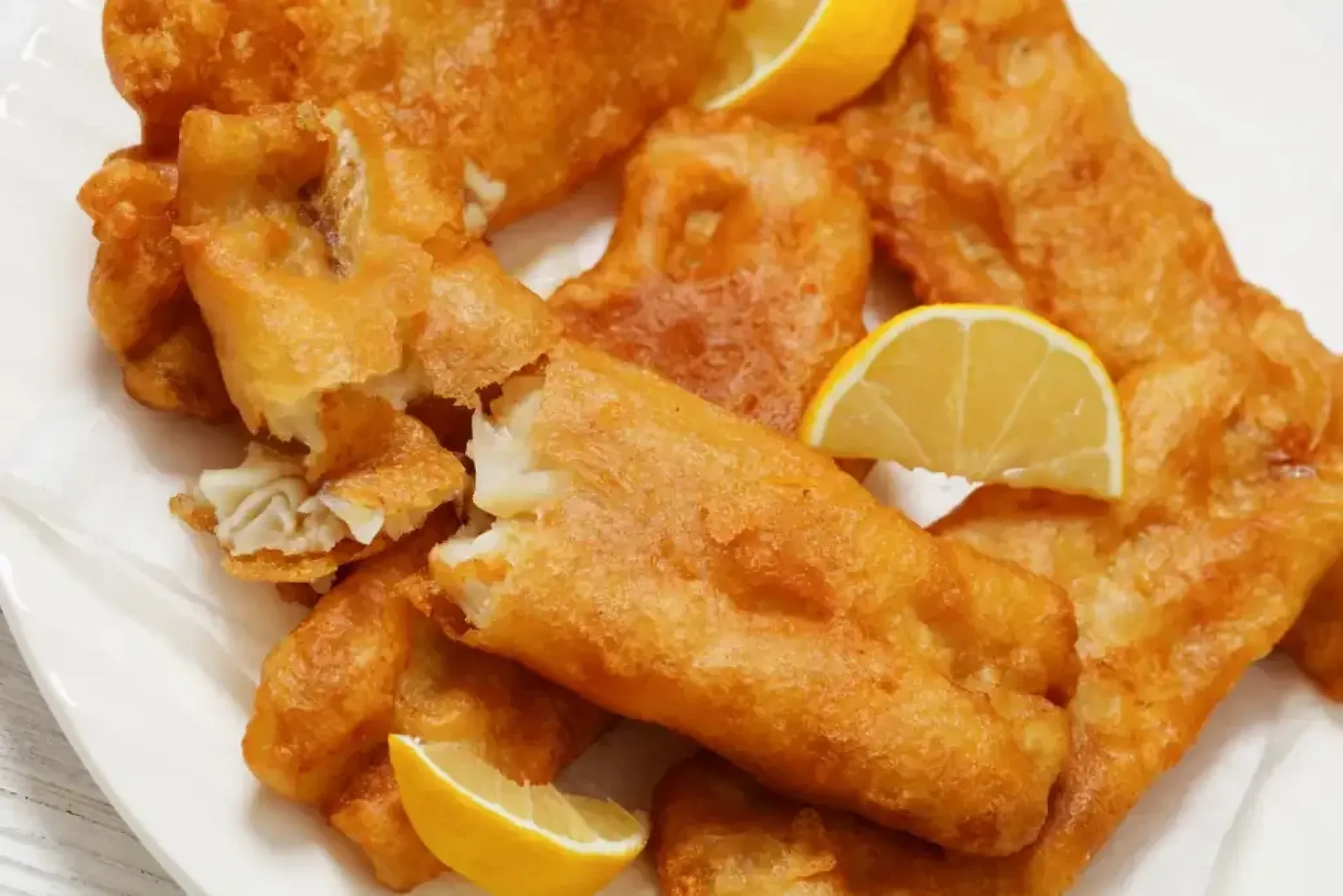 deep fried crispy beer batter fish fillet on white plate with lemon slices, dutch angle view, close-up