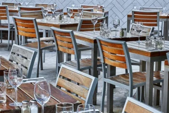 Outdoor dining area with wooden chairs featuring metal frames arranged around tables set with glasses and place settings on a patterned floor.