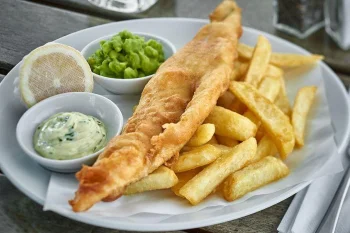 Classic fish and chips meal served on white plate with golden battered fish, thick cut chips, tartar sauce, mushy peas, and lemon wedge on wooden table