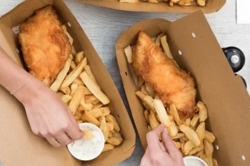 Three cardboard takeaway boxes containing battered fish and chips with white sauce cups, served on a wooden surface with hands reaching for the food.