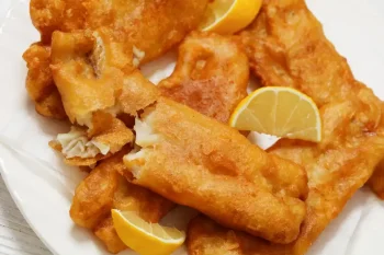 deep fried crispy beer batter fish fillet on white plate with lemon slices, dutch angle view, close-up