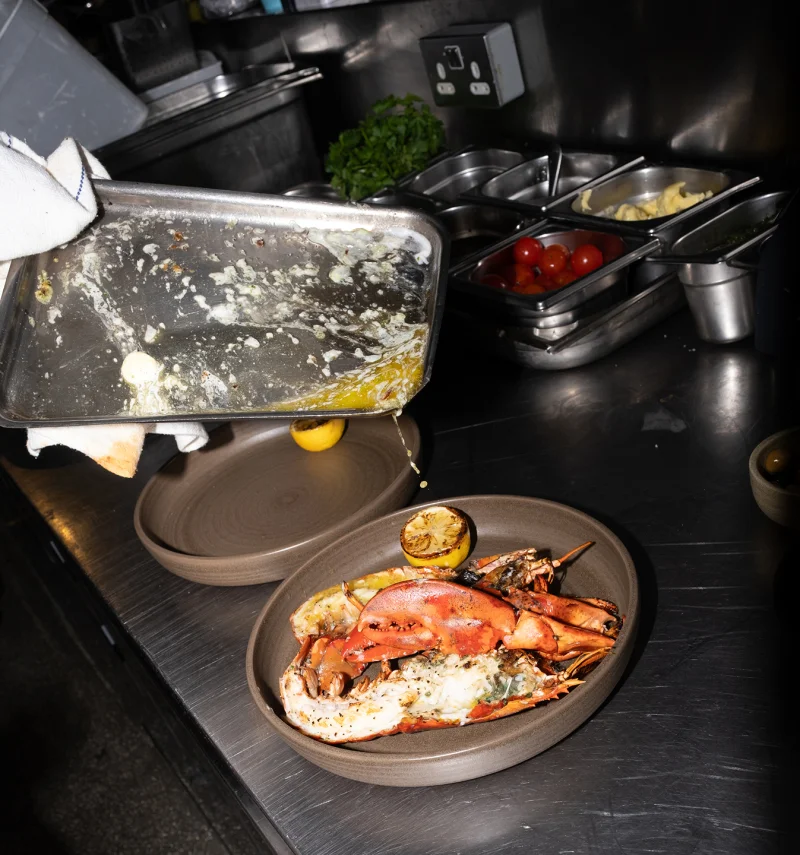 Chef plating grilled lobster with roasted lemon on a grey ceramic plate in a professional kitchen with mise en place ingredients in metal containers.