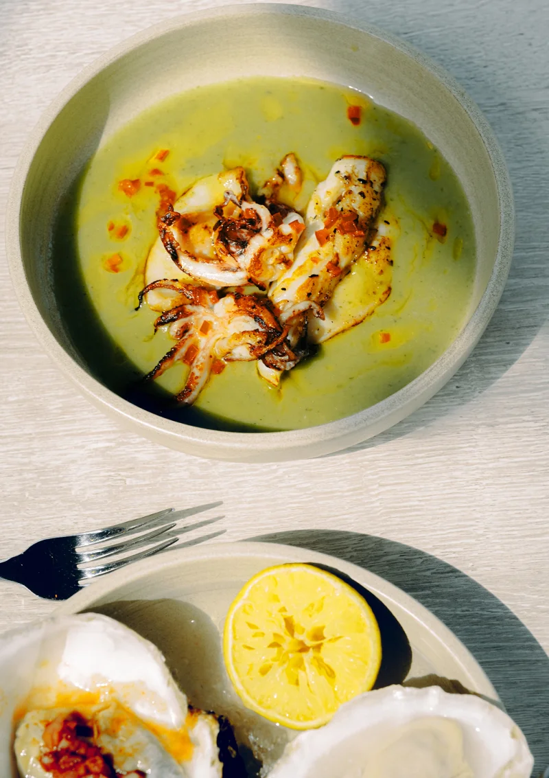 Grilled squid in green soup served in a pale bowl with a fork, accompanied by a plate with lemon halves and what appears to be eggs or seafood.