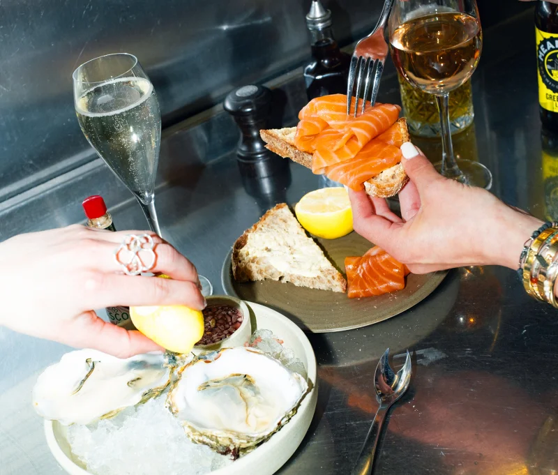 Person enjoying seafood and champagne at a bar, holding smoked salmon on toast with a fork, with oysters and lemon visible on the counter.