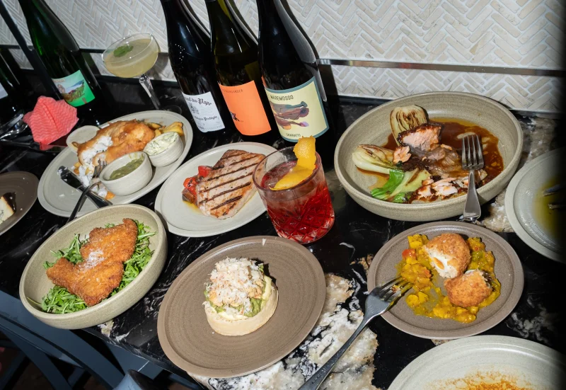 Fine dining spread with various plated dishes including seafood, meat, and vegetables, accompanied by wine bottles and a cocktail on a black table with herringbone patterned background.