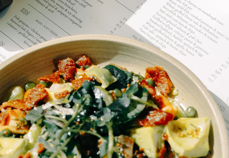Plated dish with seared seafood topped with sesame seeds, garnished with microgreens and vegetables, served on a ceramic bowl with an open menu visible in the background.