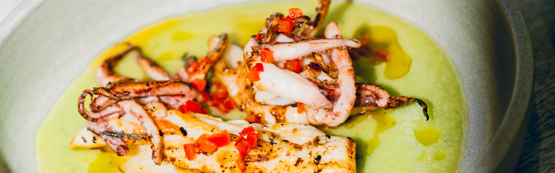 Grilled squid and fish served on a vibrant green sauce in a white bowl, garnished with red chilli, alongside additional plated dishes in the background.