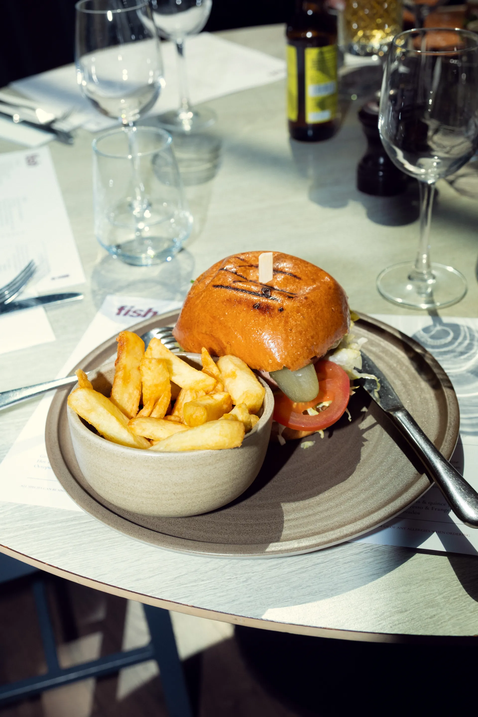 Burger with chips served in a ceramic bowl on a grey plate at a restaurant table with wine glasses and place settings