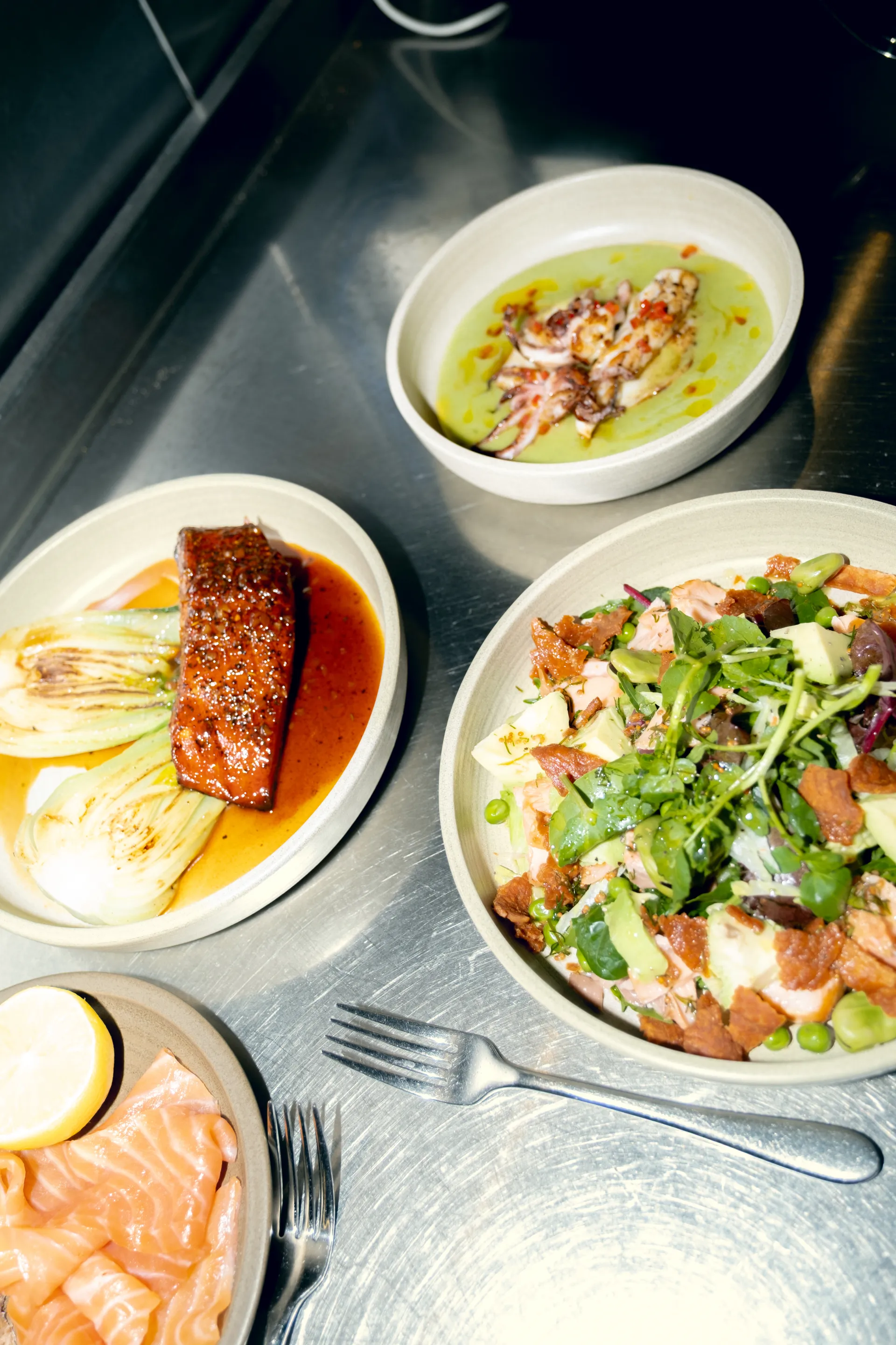 Three elegant plated dishes on a grey surface: a green soup with seafood garnish, braised meat with bok choy in brown sauce, and a salad with crispy elements and green onions, with cutlery and a glass visible.