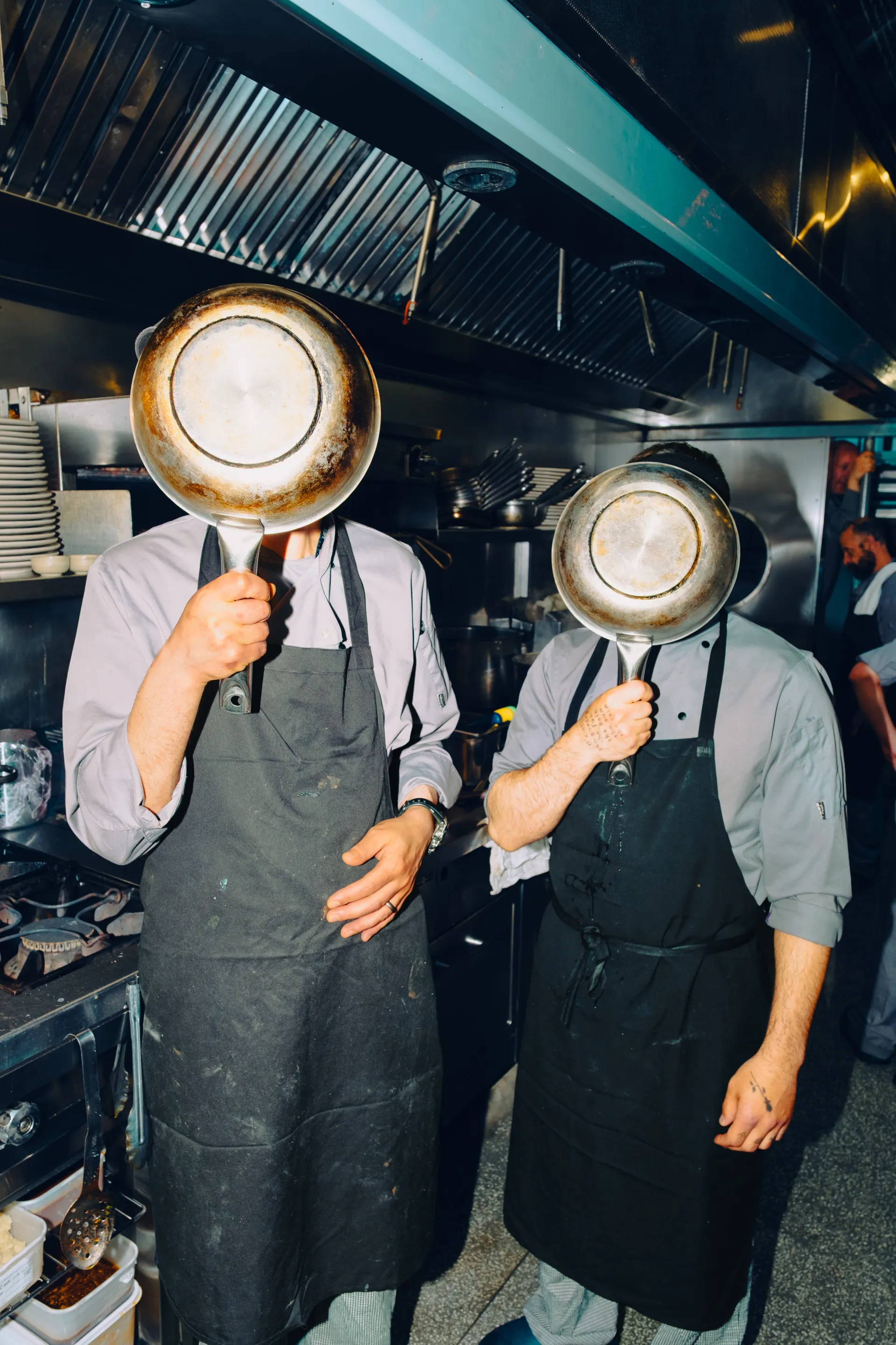 Two chefs in a kitchen holding large pans over their heads, obscuring their faces, wearing aprons and chef's whites.
