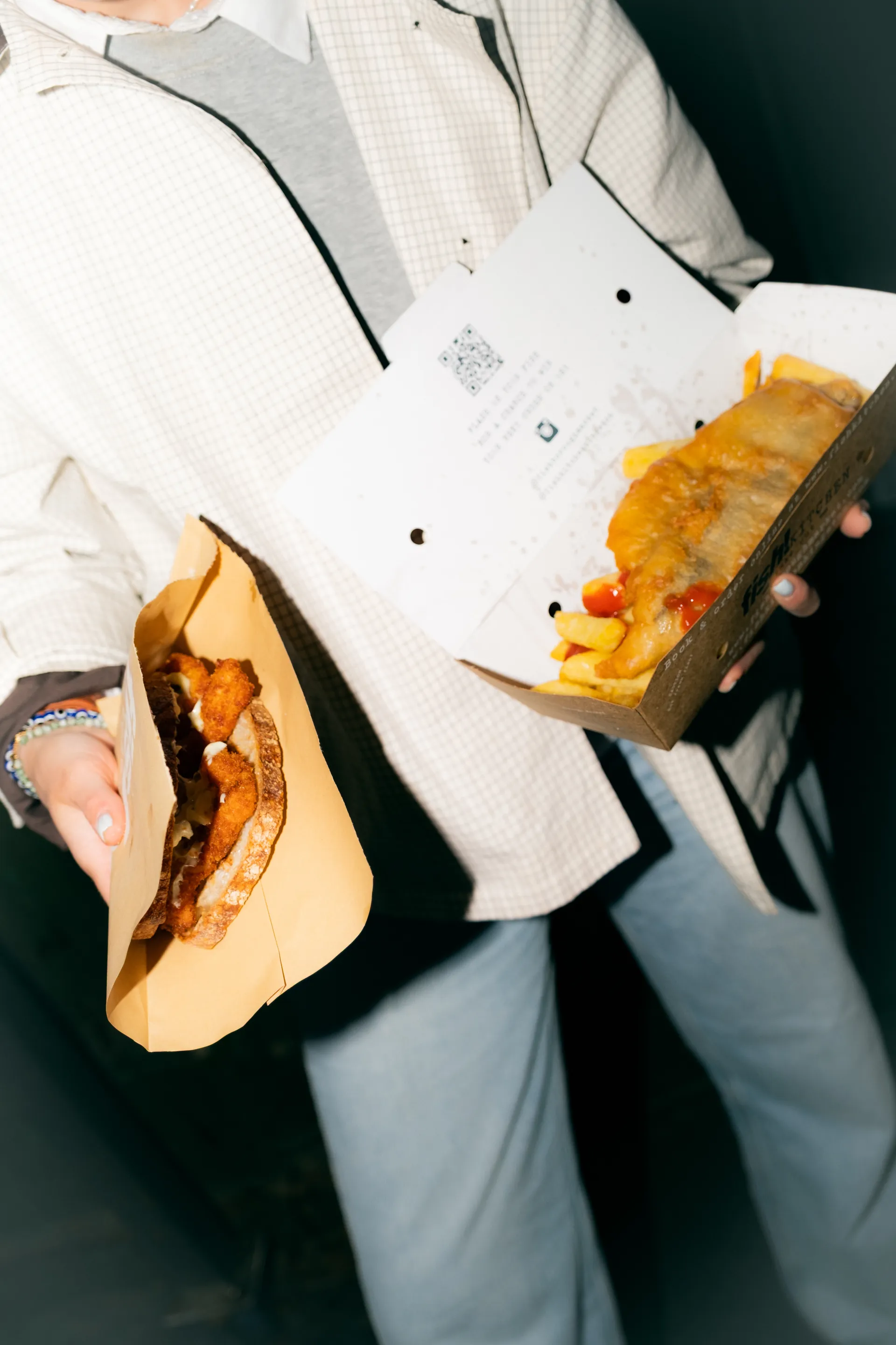 Person holding a hot dog in paper wrapping and a wooden tray of chips, wearing casual clothing against a dark background