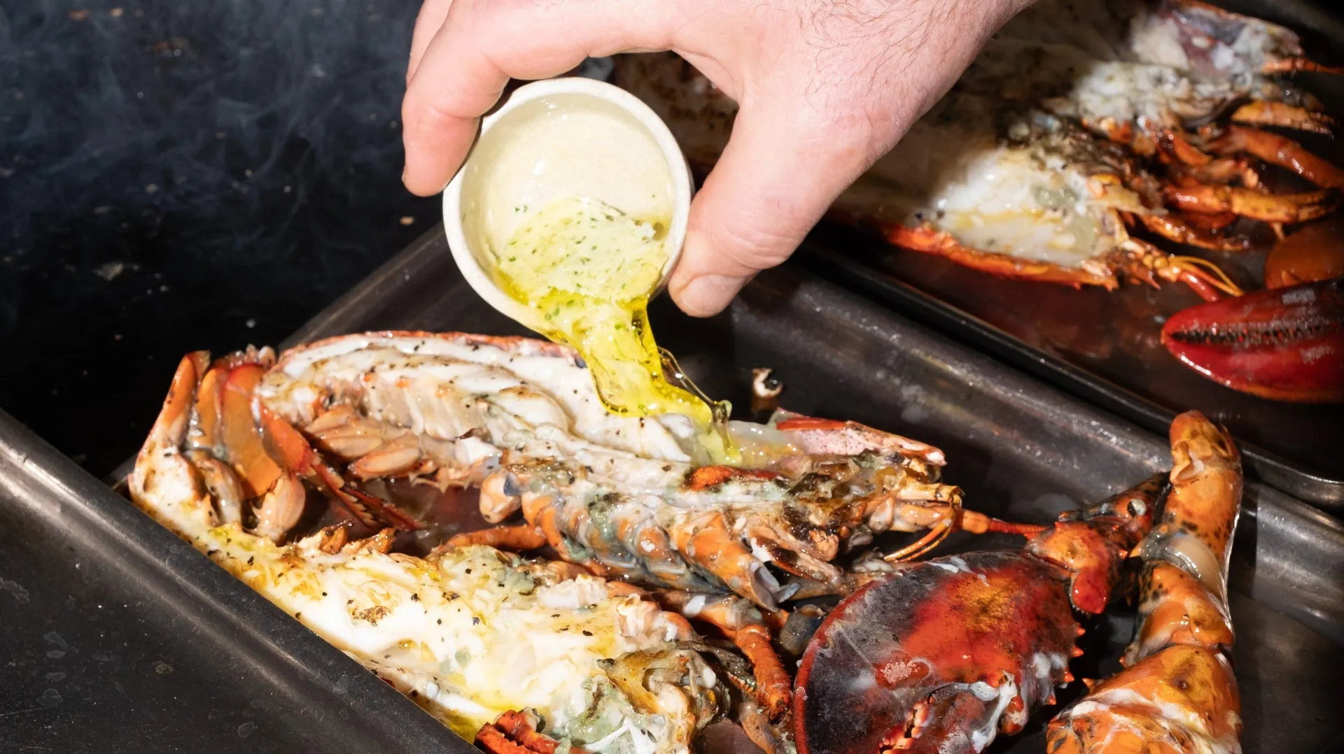 Person pouring garlic butter over cooked lobsters on a black griddle plate during a seafood cooking demonstration