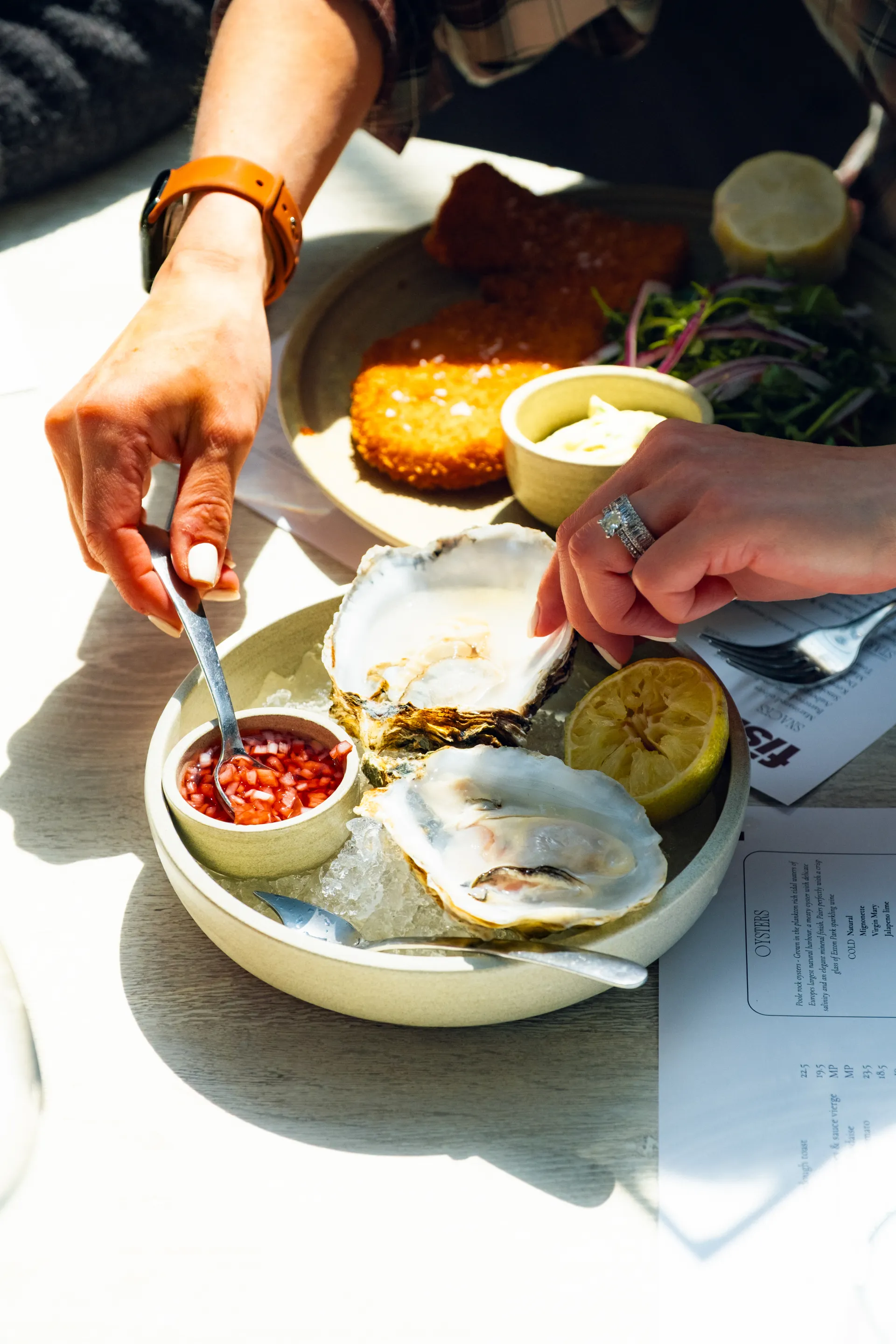 Person in sunlight eating fresh oysters with red mignonette sauce, lemon wedges, and accompaniments on a pale plate at an outdoor dining table