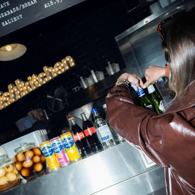 Person in brown leather jacket selecting a bottle from a bar counter displaying various beverages and snacks, with illuminated signage visible in the background.