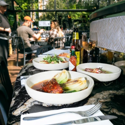 Outdoor dining scene with plated dishes featuring glazed meat and vegetables, beer bottles, wine glass, and diners in background under green umbrellas.