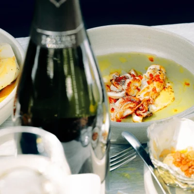 Elegant seafood dish with seared scallops in butter sauce, garnished with red chilli, served alongside champagne and white plates against dark background.