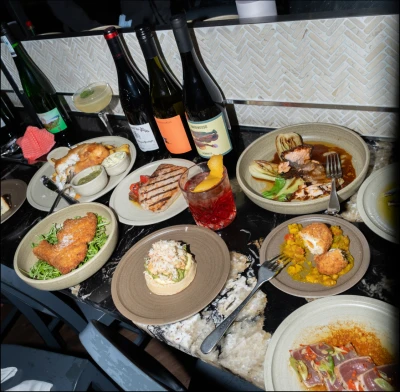Fine dining spread with multiple gourmet dishes including seafood, meat, and vegetables served on beige plates, accompanied by wine bottles and cocktails against a herringbone patterned backdrop.