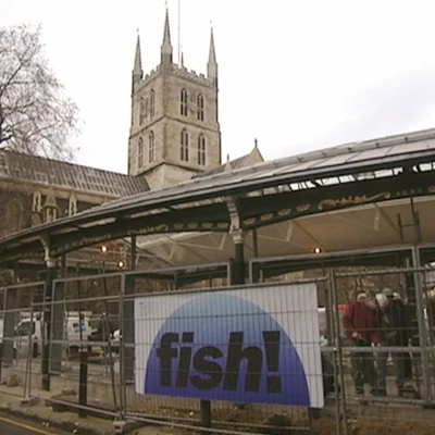 A stone church with a tall spire stands behind a modern market structure with a blue "Lisha" logo sign displayed on metal fencing.