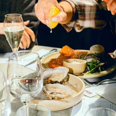 Fine dining table setting with fresh oysters, champagne flutes, roasted poultry, and a person squeezing lemon over seafood during an elegant meal
