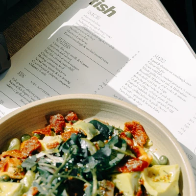 Restaurant menu displayed above a ceramic bowl containing seared seafood with green vegetables and potatoes on a wooden table surface