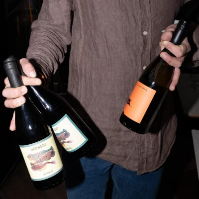 Person in grey shirt holding two wine bottles - Maremosso Carignatto on the left and an orange-labelled bottle on the right, against dark background.