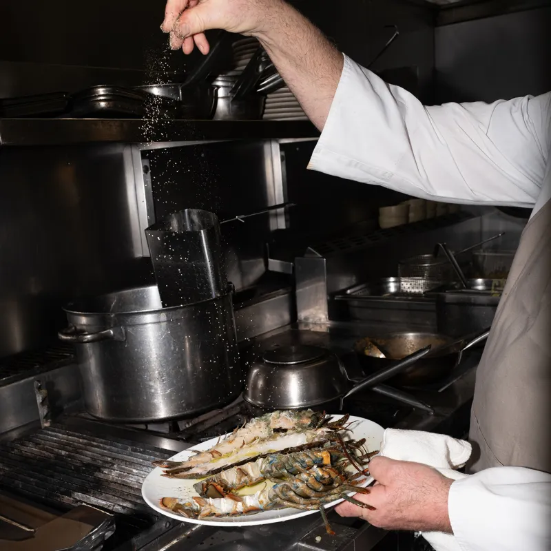 Chef in white chef's coat seasoning grilled lobsters on a plate in a professional kitchen with pots and pans on the stove.